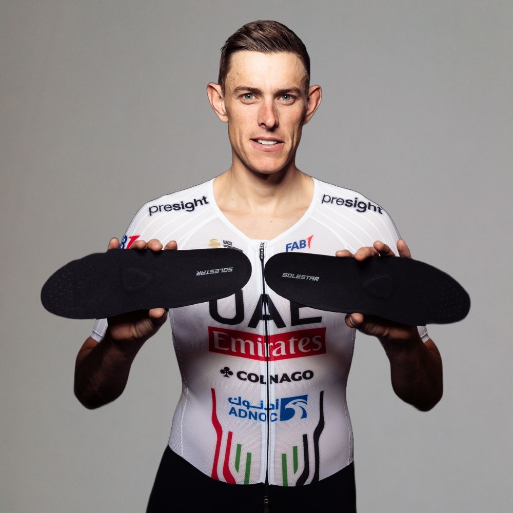 Person wearing a cycling jersey holding two black insoles against a gray background