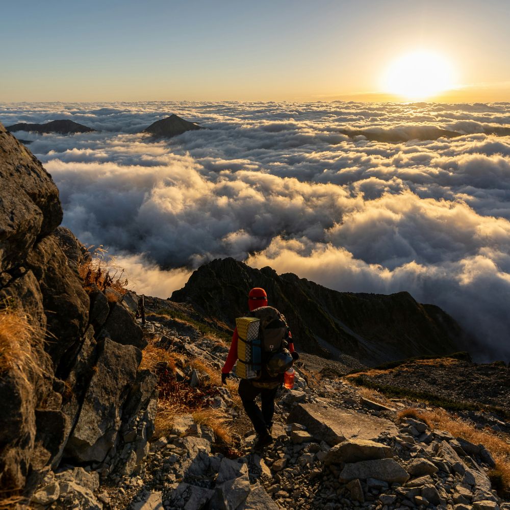 Person hiking on a mountain ridge with a scenic view of clouds below at sunset.