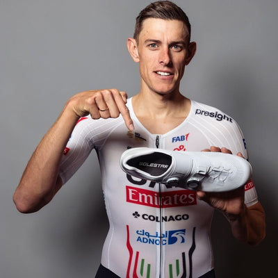 Cyclist Nils Pollitt holding a cycling shoe with sponsor logos on a gray background